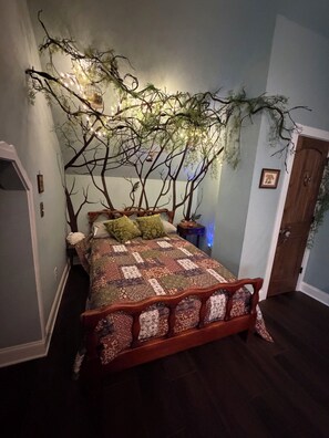 Een bed met een gebedekt patchworkdeken, een houten hoofdbord en een aan de muur bevestigde decoratie van een boom met lichtjes.