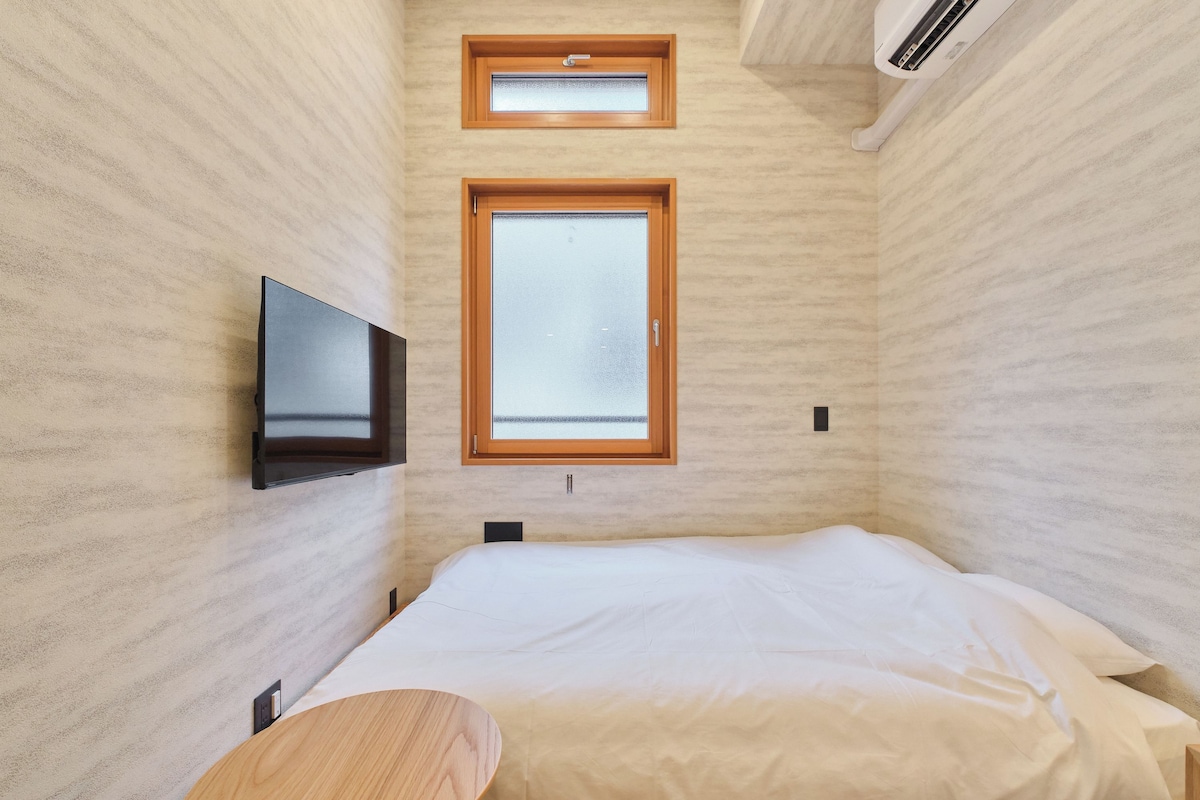 A bedroom with a bed, a television mounted on the wall, and two windows.