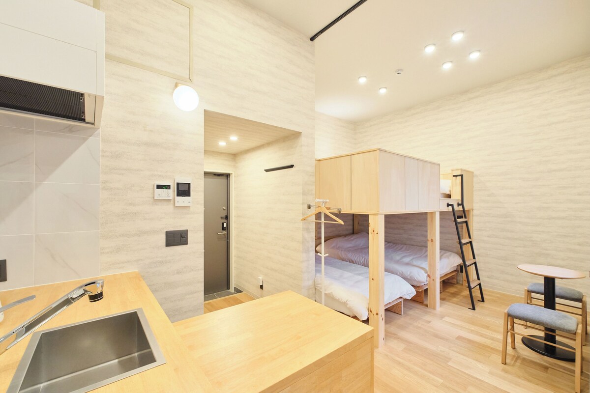 A compact hotel room with a bunk bed, a small kitchenette, a single table, and a chair.