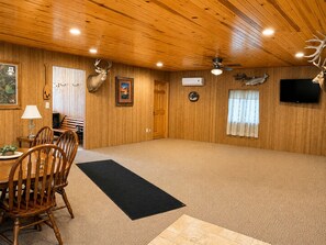A spacious room with wooden walls, a ceiling fan, a mounted television, and a dining area with a table and chairs.
