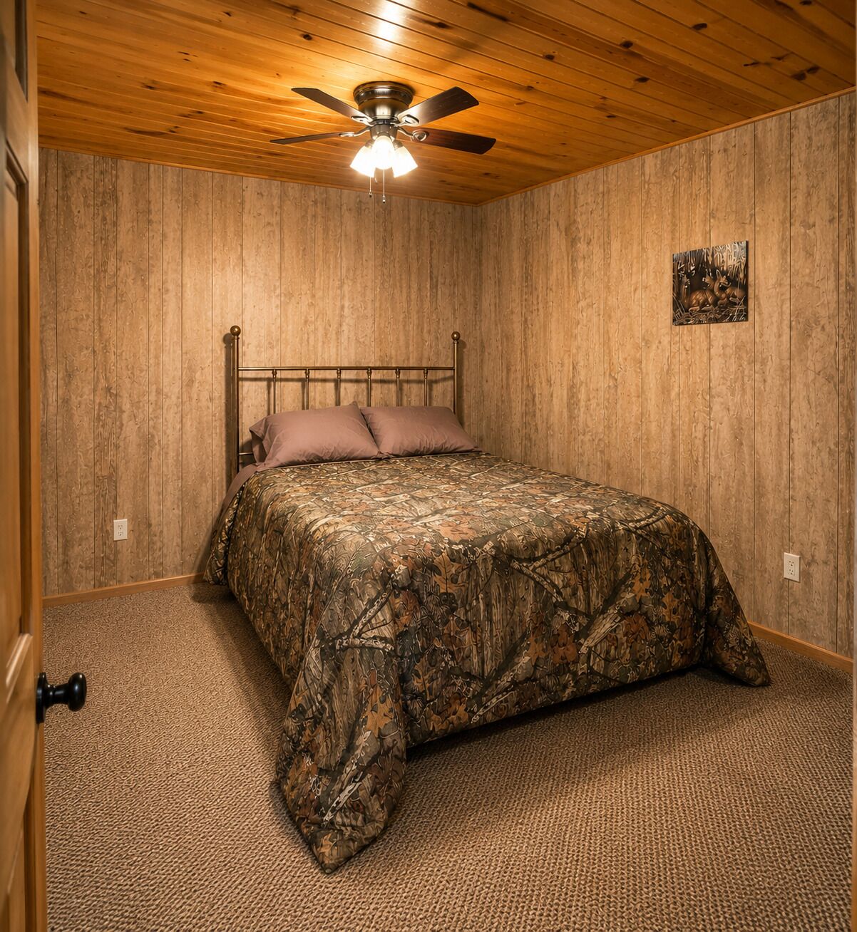 Una camera da letto con soffitto in legno, un letto con copriletto mimetico, una ventola a soffitto e un piccolo quadro appeso al muro.