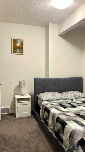 A bedroom with a bed, a nightstand, a lamp, and a painting on the wall. - Kennis Home (Airdrie)