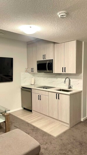 A compact kitchen with light wood cabinets, a microwave, a sink, and a dishwasher. - Kennis Home (Airdrie)