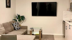A living room with a sofa, a glass coffee table, a TV mounted on the wall, and a kitchen area with cabinets. - Kennis Home (Airdrie)
