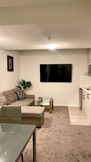 A living room with a sofa, a glass coffee table, a TV mounted on the wall, and a kitchen area with cabinets. - Kennis Home (Airdrie)