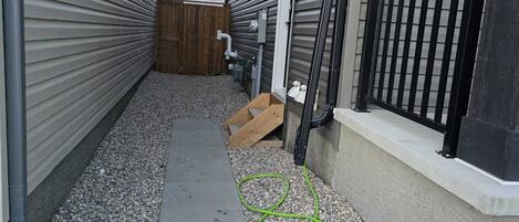 Een smal betonnen pad dat leidt naar een houten terras met een metalen hek, omringd door huizen met bekleding en een zwart metalen omheining.