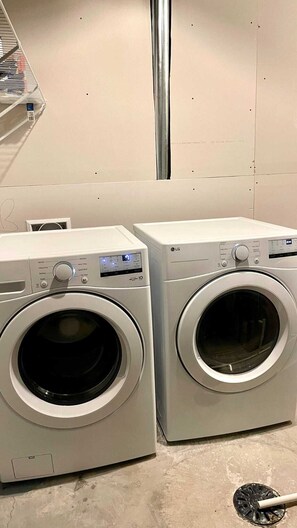 Two front-loading washing machines in a laundry room with a metal vent and a shelf with laundry supplies. - Kennis Home (Airdrie)