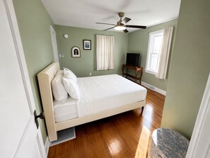 A bedroom with a bed, a ceiling fan, a television on a stand, and a window with curtains.