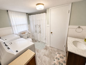 Een badkamer met een wit toilet, een wastafel met een marmeren blad, een douche met een bloemen gordijn en een wasmachine.