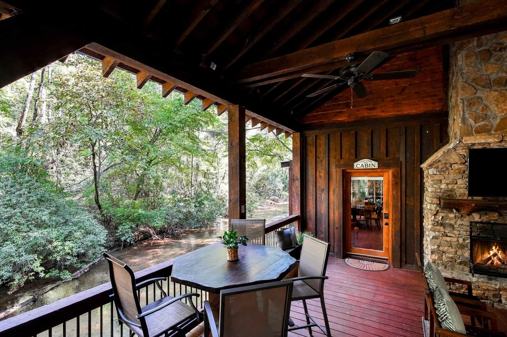 A wooden deck with a table and chairs, a stone fireplace, and a view of a forested area.