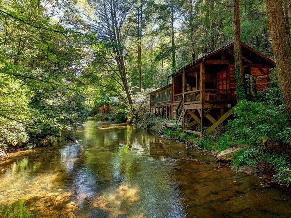 Una cabaña de madera con terraza sobre un río, rodeada de un bosque denso.