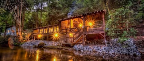 A wooden cabin with a deck and a stone pathway leading to a reflective water body.
