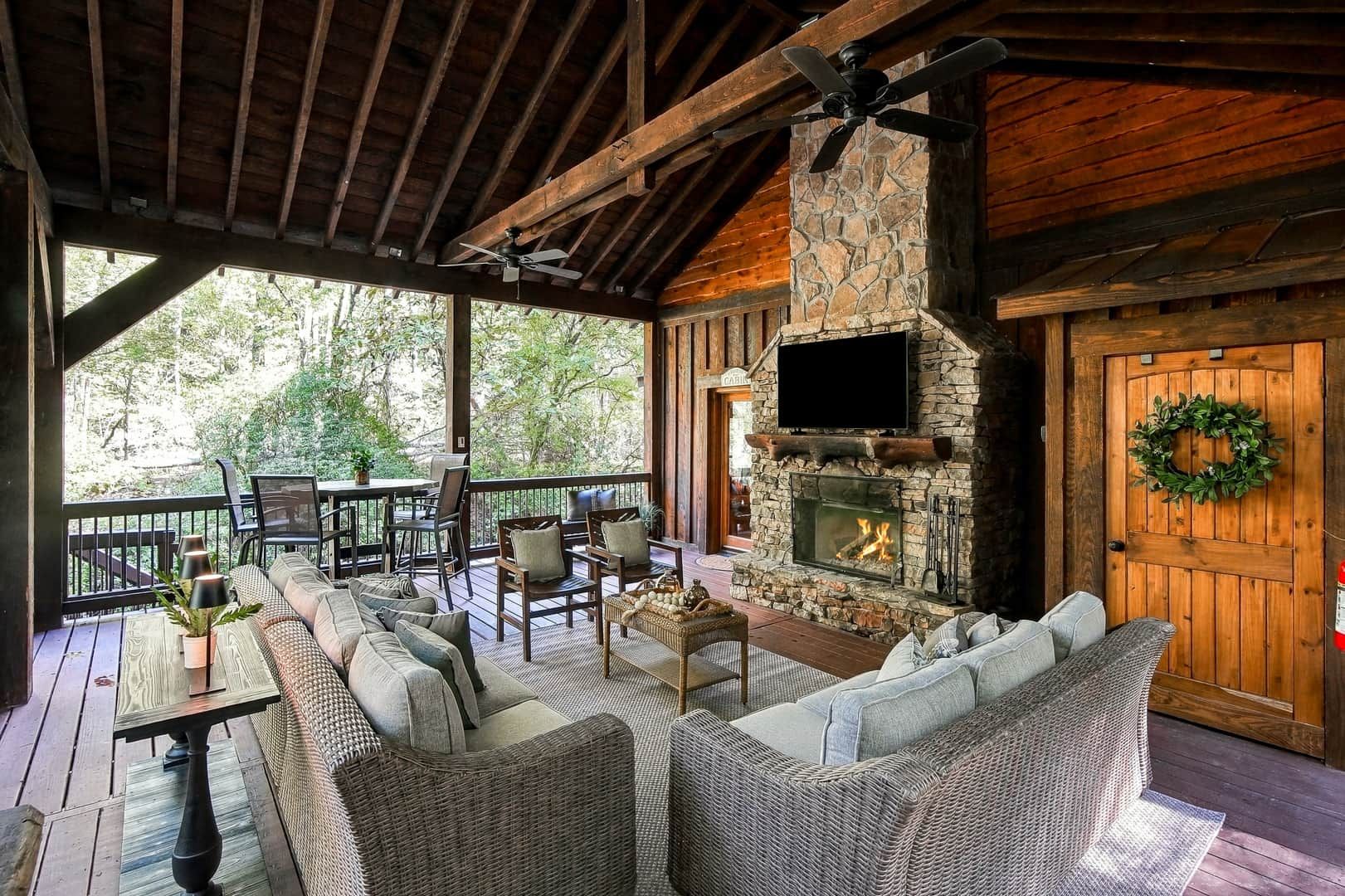 A covered outdoor seating area with wicker furniture, a stone fireplace, and a television mounted on the wall.