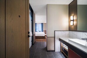 Habitación de hotel con puerta de madera, cama, baño con lavabo y espejo, y vista al pasillo.