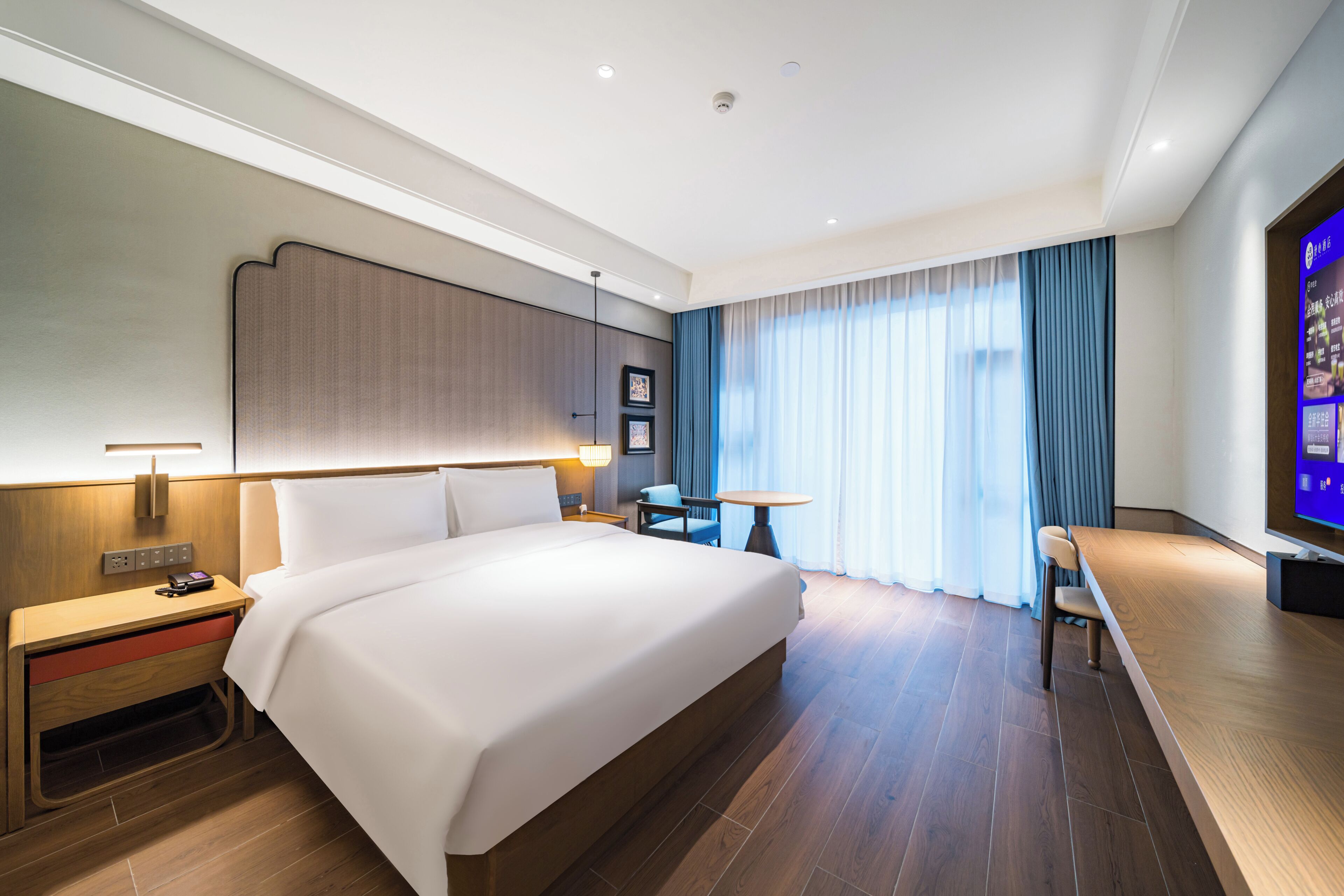 A hotel room with a large bed, bedside tables, a desk with a chair, a television, and a window with curtains.