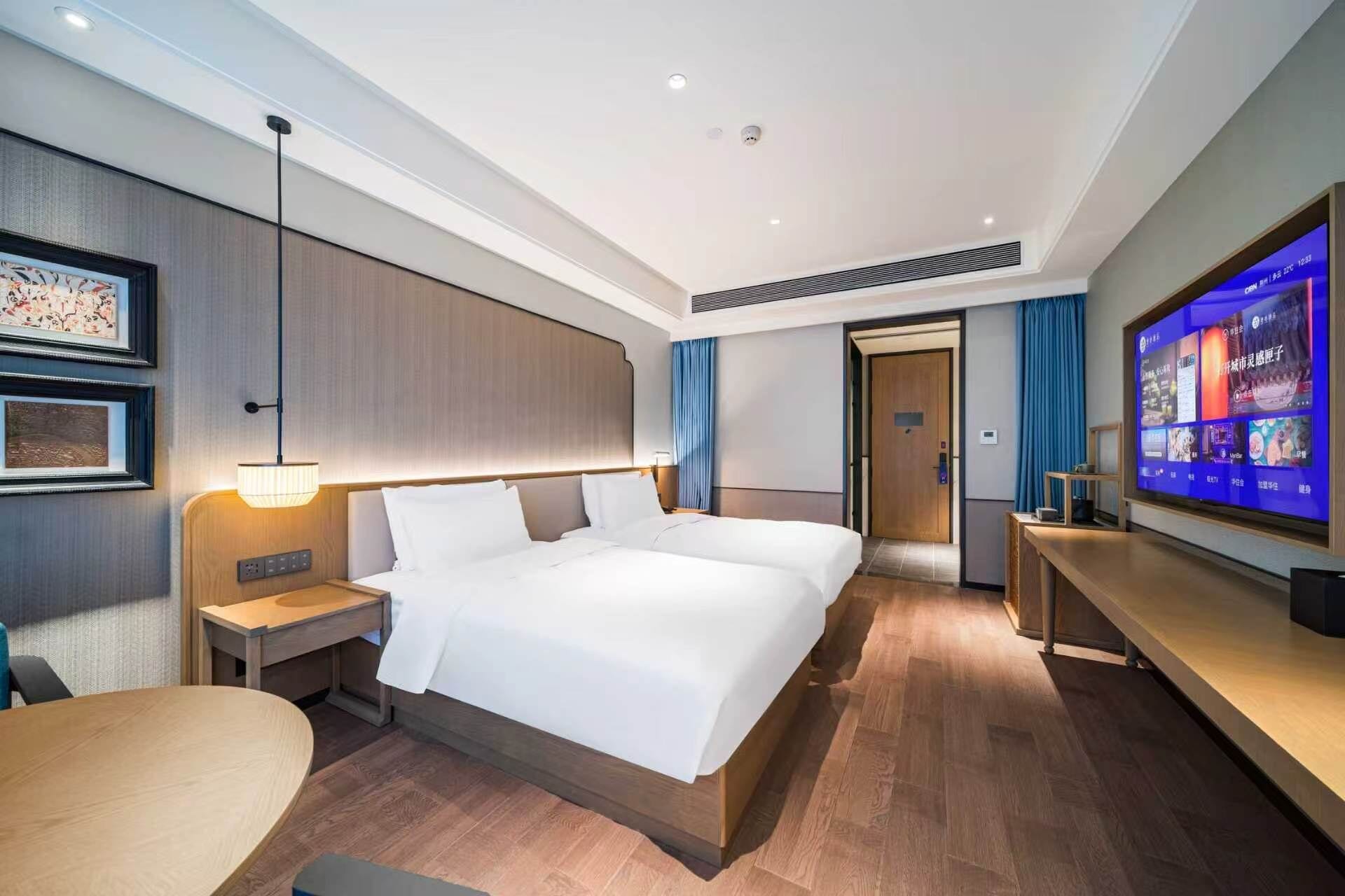 A modern hotel room with a large bed, bedside tables, a desk, a flat-screen TV, and a ceiling with recessed lighting.