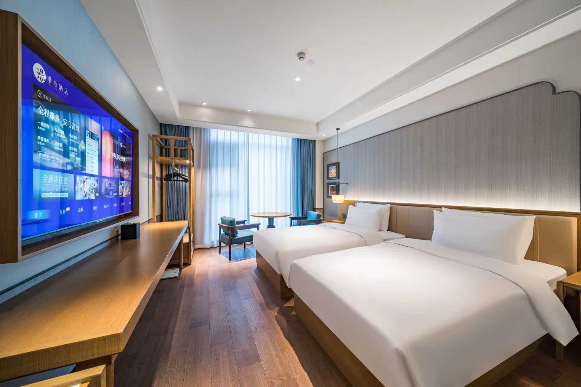 A hotel room with a large flat-screen TV, a desk, two beds, a small table, and a chair.