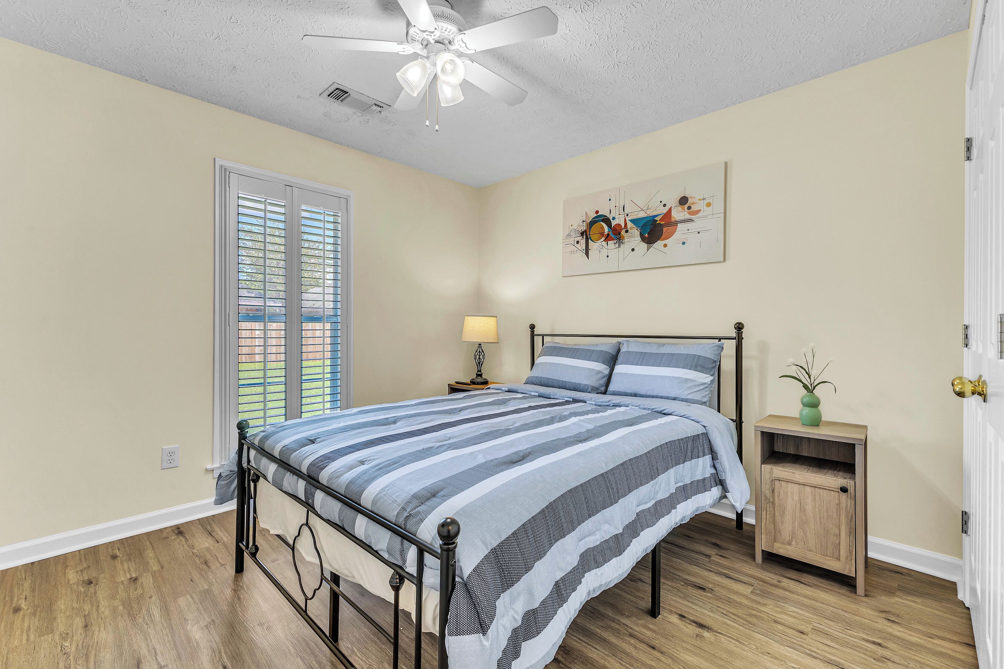 A bedroom with a bed, a nightstand, a lamp, a ceiling fan, a window with blinds, and a door.