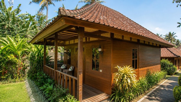 Ein Holzbungalow mit Ziegeldach, einer Veranda mit Sitzgelegenheit und üppiger tropischer Vegetation.