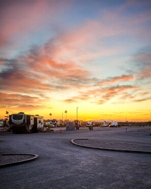 A parking area with RVs and trailers, surrounded by a desert landscape with a sunset sky.