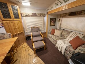 A compact RV interior with a wooden table, a cushioned chair, a sofa, and a window with blinds. - Cozy 1-bedroom recreational vehicle with private beach accesses site included. (Ehrenberg)