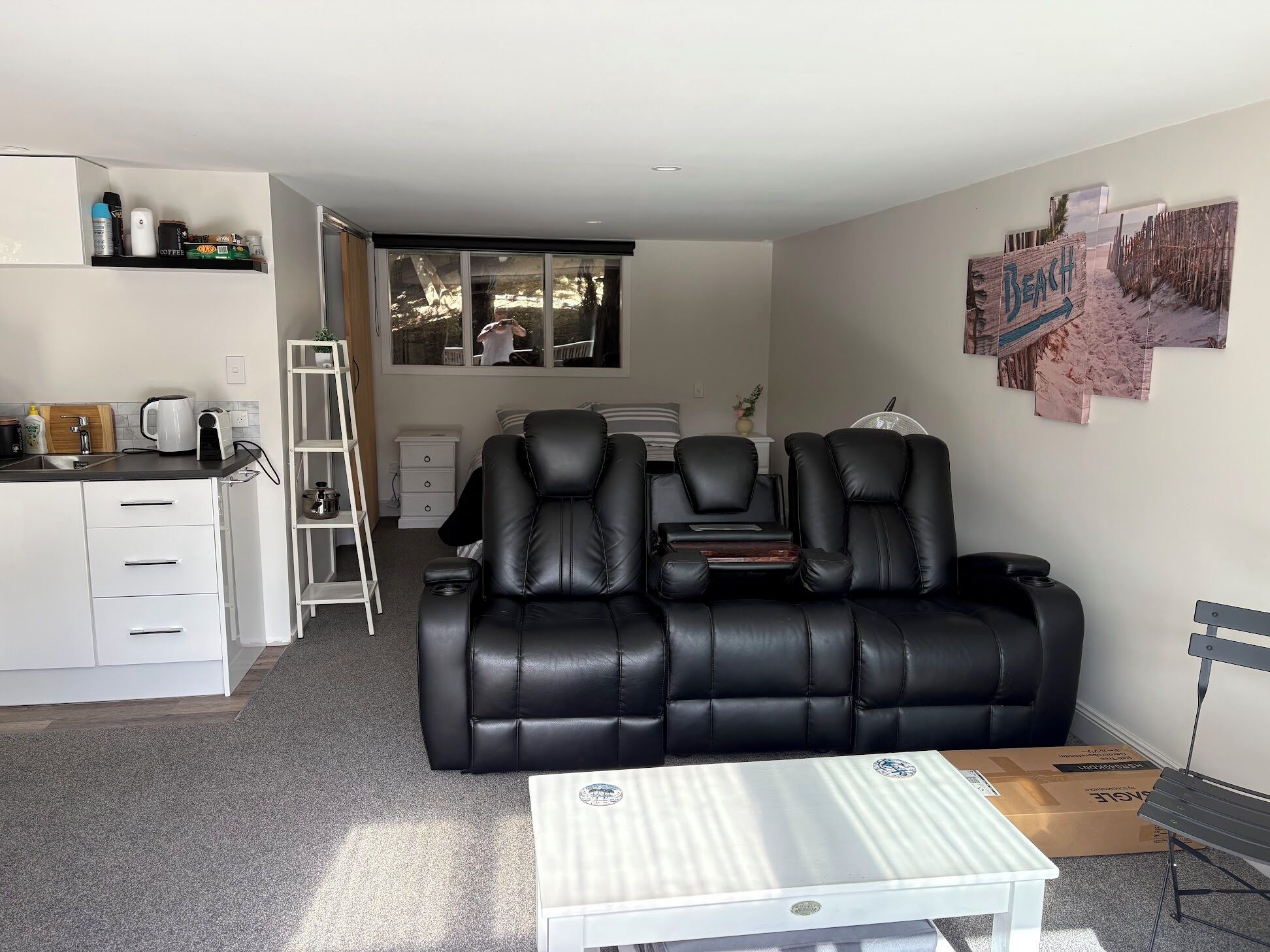 A modern living room with a black leather sofa, a white coffee table, and a kitchen area with a kettle and coffee maker.