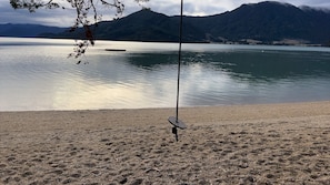 A beach with a swing set and a calm lake in the background. - Perfect Marlborough Sounds cabin retreat for your Sandy Bay getaway (Black Rock)
