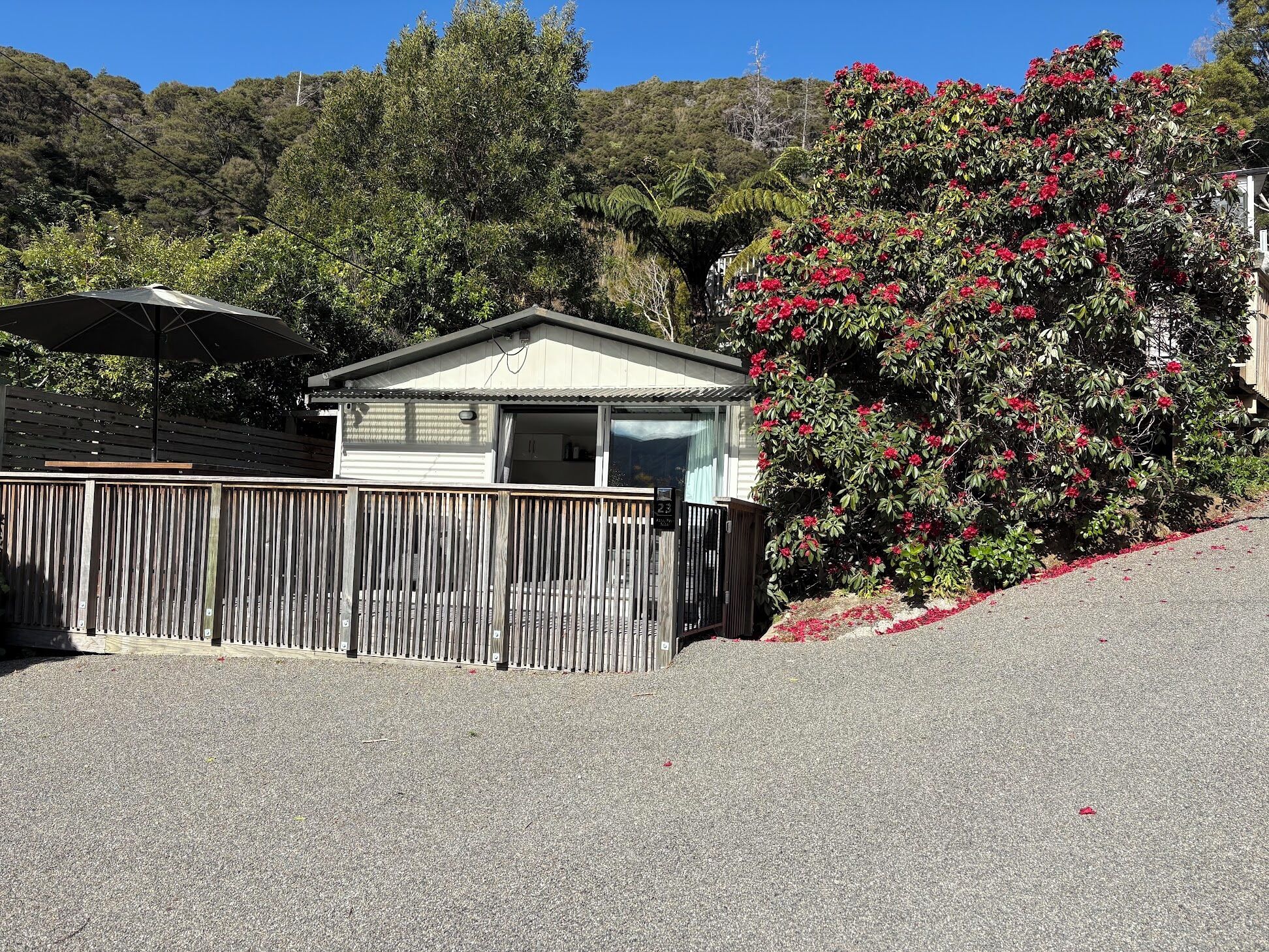 A small, single-story building with a flat roof, a wooden fence, and a gravel driveway.