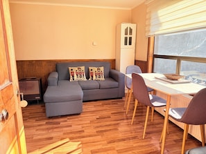 A room with a grey sofa, a dining table with chairs, a radiator, and a window with blinds.