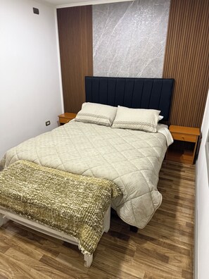 A neatly made bed with a quilted headboard, a bedside table, and a wooden floor.