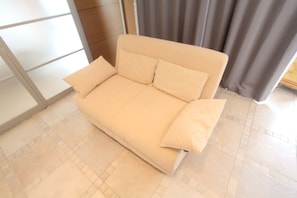 A beige sofa with two pillows in a room with tiled flooring and a window with blinds. - Spacious Private Stay Near Higashi-Shinjuku | Sleeps 10 | Families & Groups | WiFi (2) (新宿区新宿７丁目)