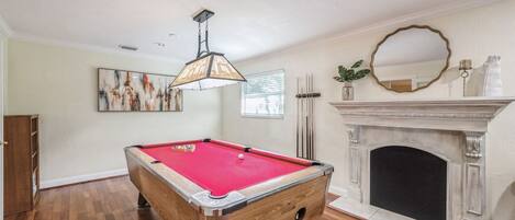 A pool table in a room with a fireplace, a painting on the wall, and a ceiling light fixture.