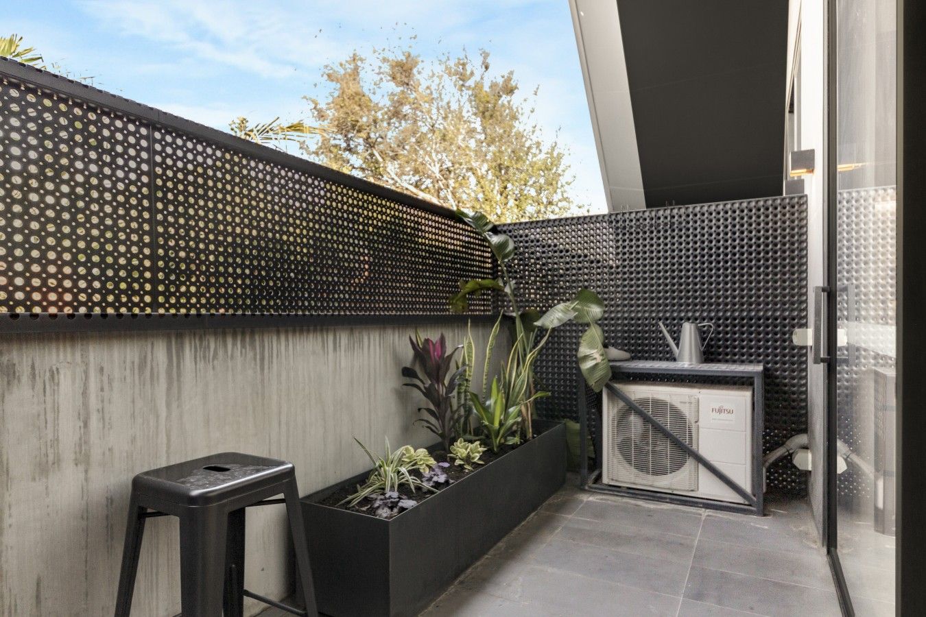 Um espaço ao ar livre moderno com uma parede preta perfurada, um vaso de concreto com plantas, um banquinho preto e um aparelho de ar condicionado branco.