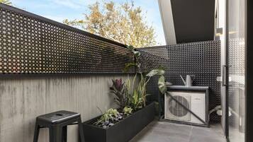 A modern outdoor space with a black perforated wall, a concrete planter with plants, a black stool, and a white air conditioning unit.