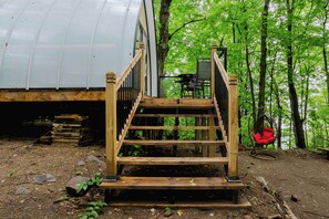 Un escalier en bois mène à une terrasse aménagée avec du mobilier d’extérieur, le tout étant entouré d’une forêt.