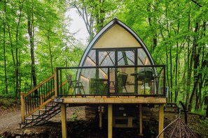 Une petite cabane triangulaire en bois, dotée d’un balcon en verre et entourée d’une forêt.