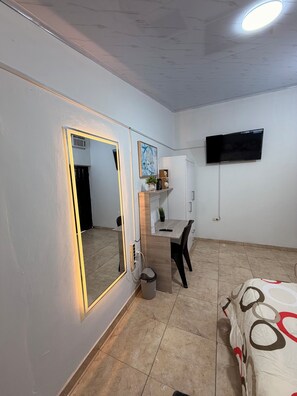 A hotel room with a bed, a desk, a chair, a TV mounted on the wall, a mirror with a light, and a trash bin.
