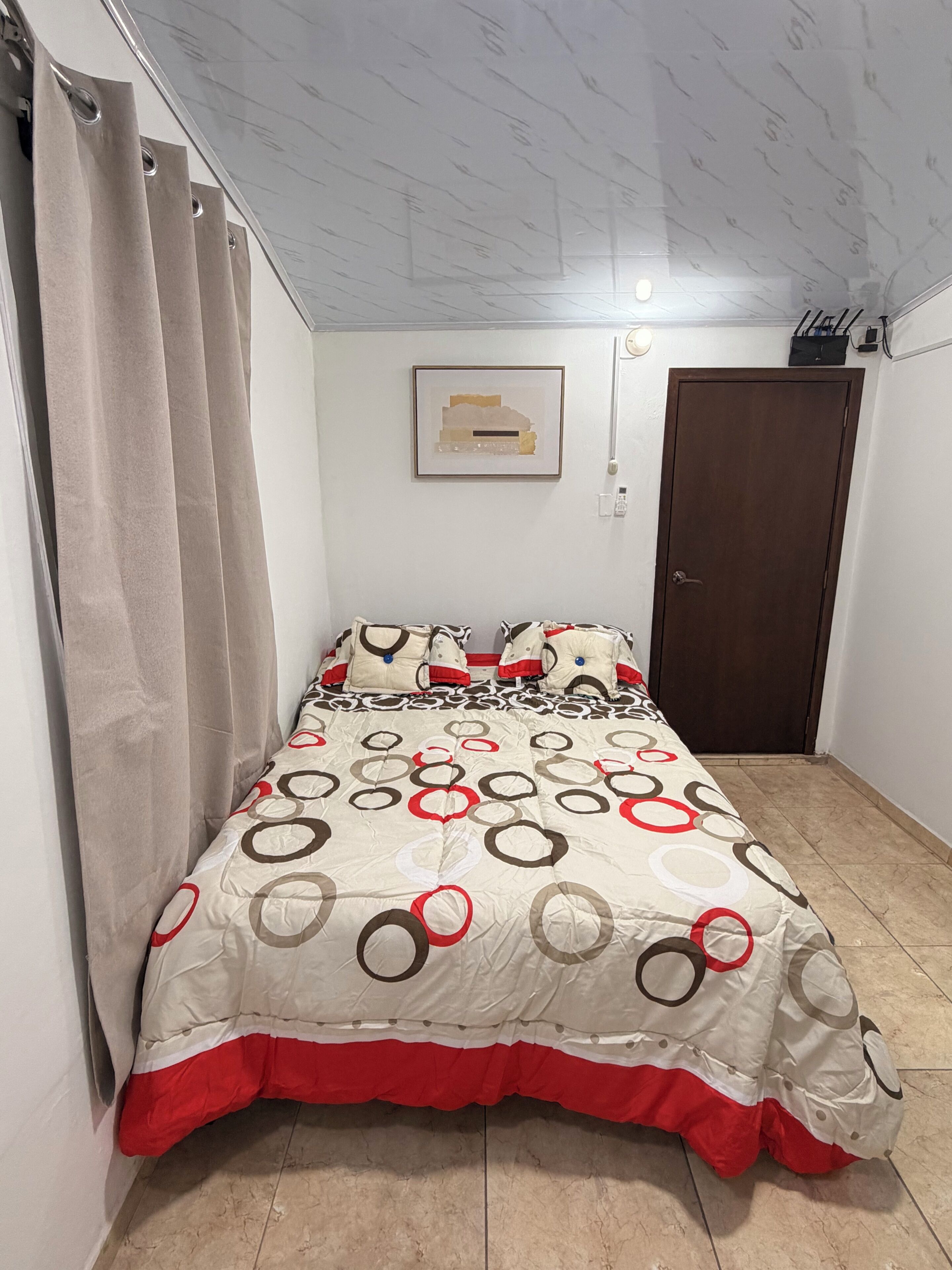 Un dormitorio con una cama, cortinas, una puerta, una lámpara fijada en la pared y un cuadro enmarcado.