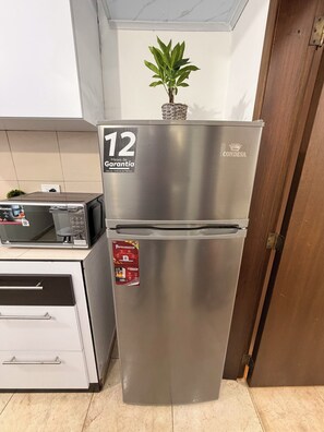 A stainless steel refrigerator with a 12-month warranty label, a small potted plant on top, and a microwave on the left.