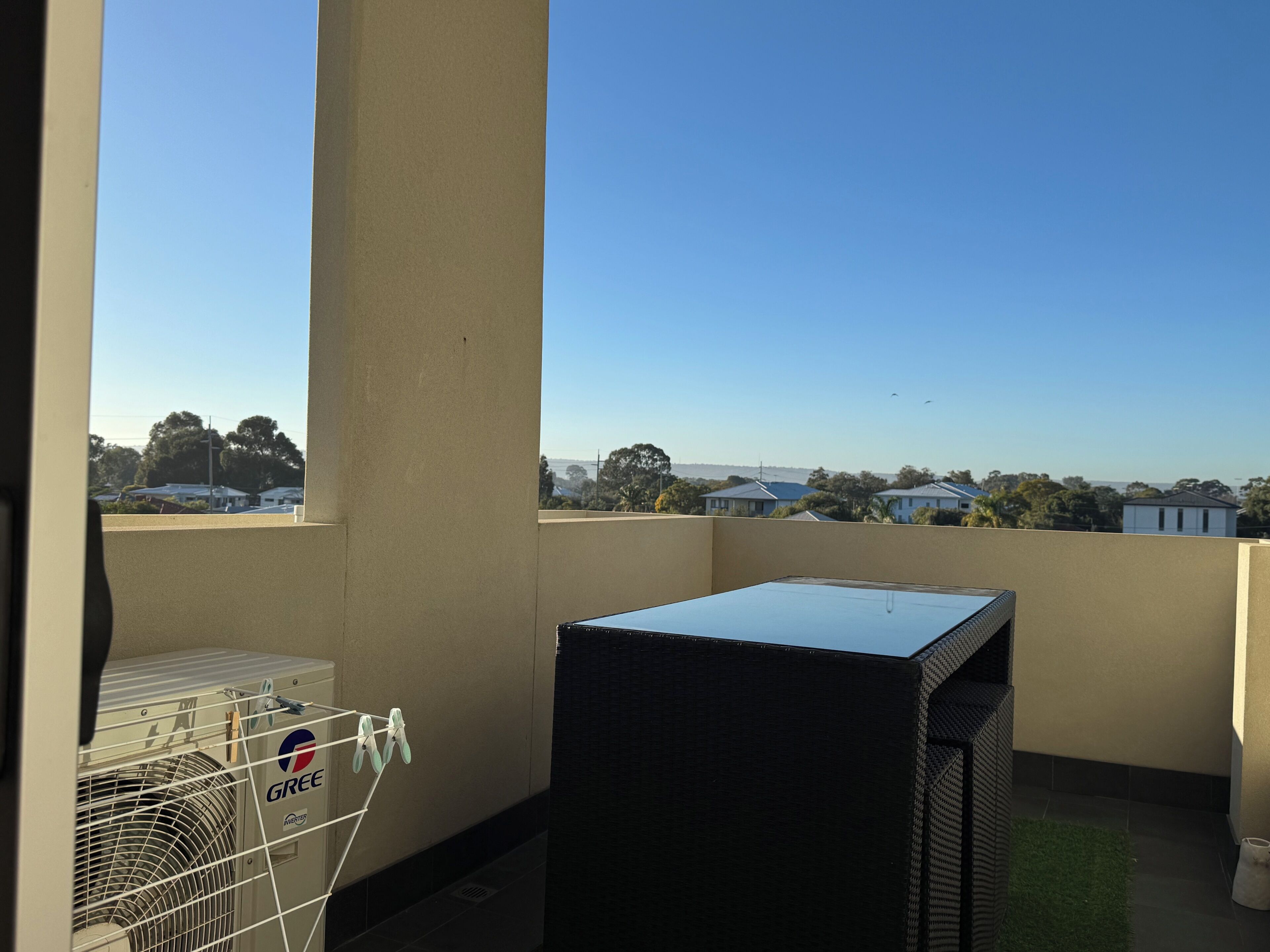 A balcony with a view of houses and trees, a black grill, and a white laundry basket.