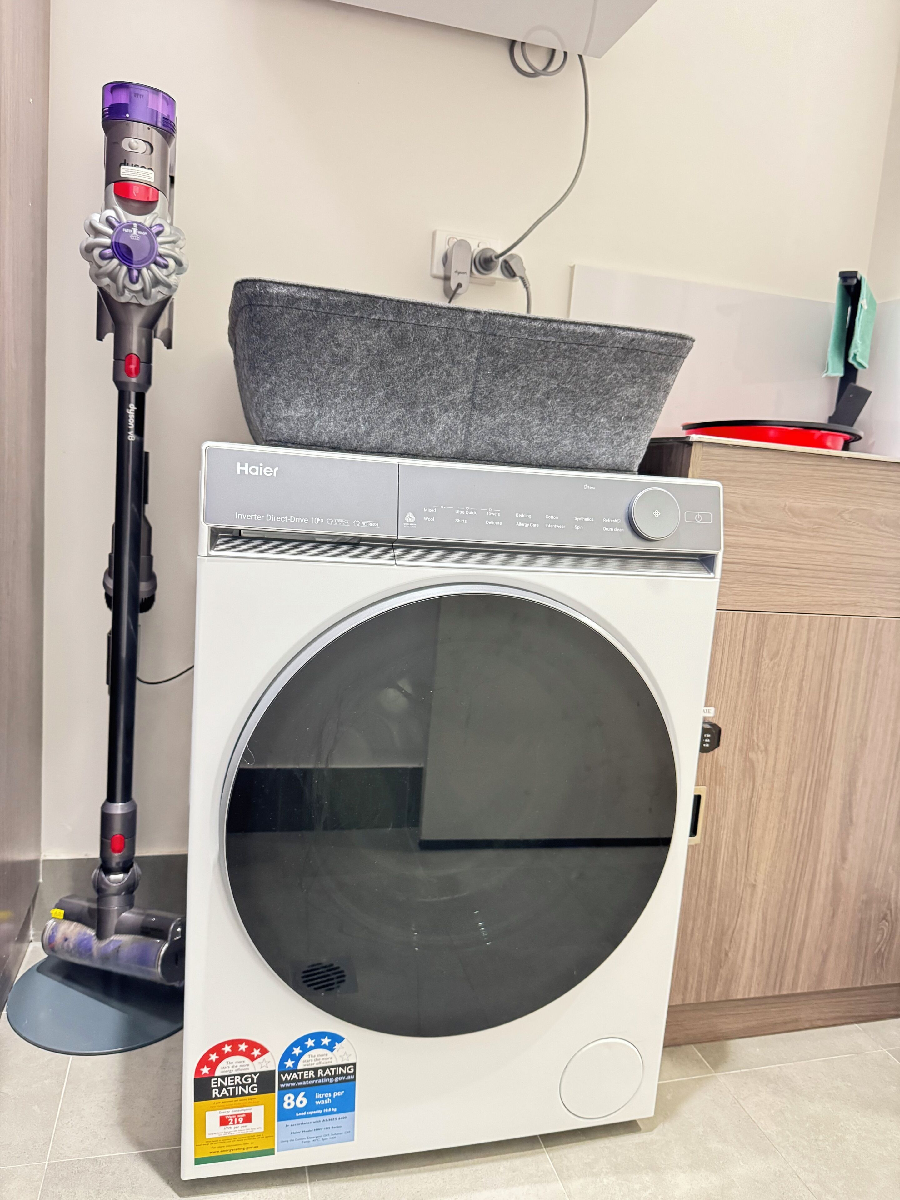 A front-loading washing machine with a control panel, a vacuum cleaner, and a kitchen area with a red pot on the counter.