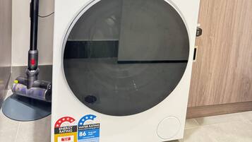 A front-loading washing machine with a control panel, a vacuum cleaner, and a kitchen area with a red pot on the counter.
