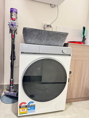 A front-loading washing machine with a control panel, a vacuum cleaner, and a kitchen area with a red pot on the counter. - Blissful Modern Apartment (Cloverdale)