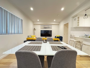 Een moderne woonkamer met een eettafel, grijze stoelen, een bank met gele kussens, een flatscreen televisie en een keuken met een mozaïekachtergrond.
