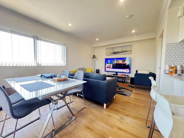 Een moderne woonkamer met een glazen eettafel, een grijze bank en een flatscreen televisie waarop nieuws wordt vertoond.