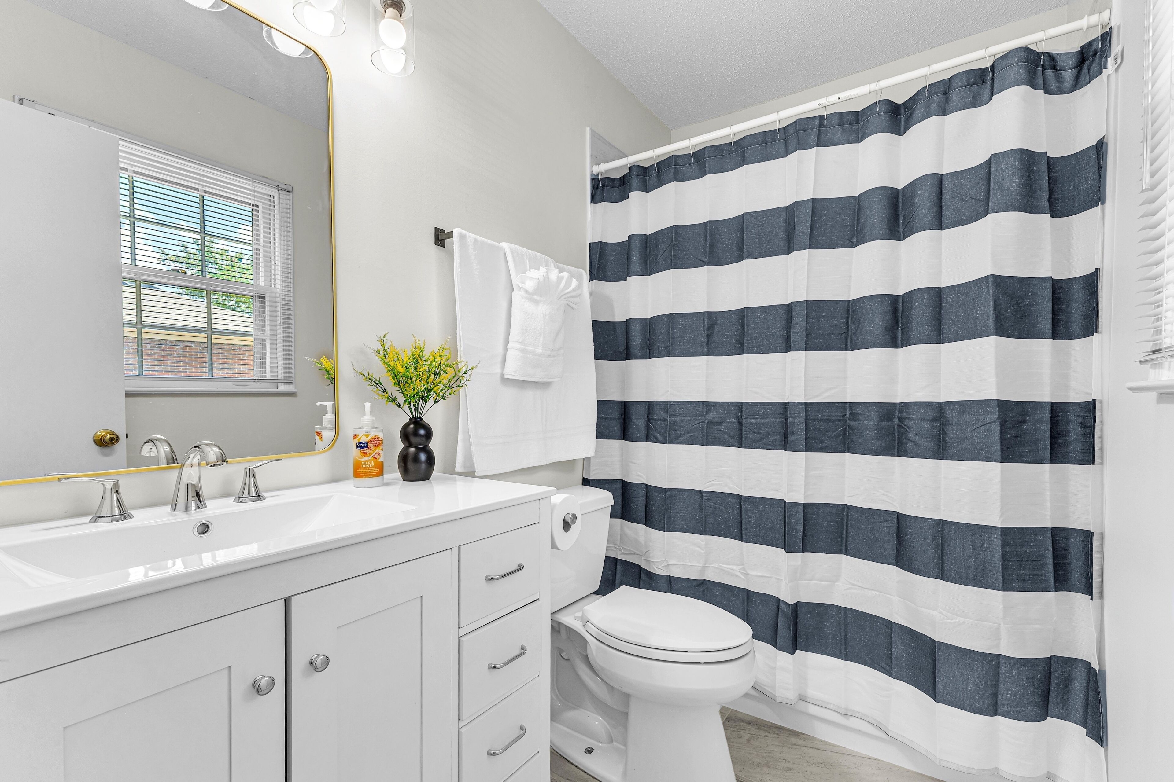 Une salle de bain avec un lavabo blanc, deux robinets, un miroir avec éclairage intégré, des toilettes et un rideau de douche à rayures.