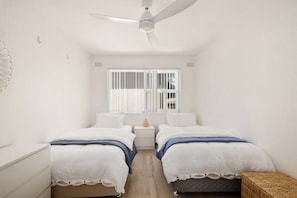 A bedroom with two beds, a ceiling fan, a window with blinds, and a bedside table with a lamp. - Kosynook - On the Beach (Mona Vale)