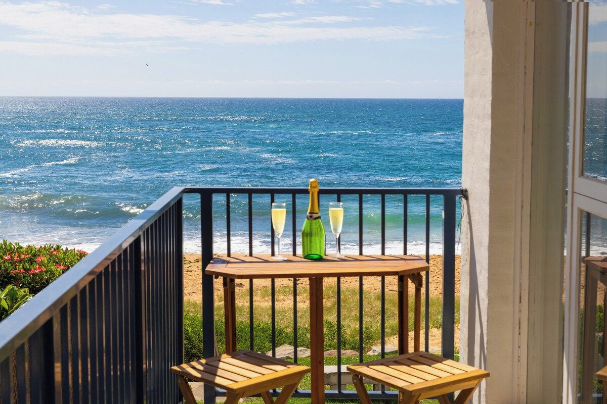 A balcony with a view of the ocean, a wooden table, two chairs, and two glasses of champagne.