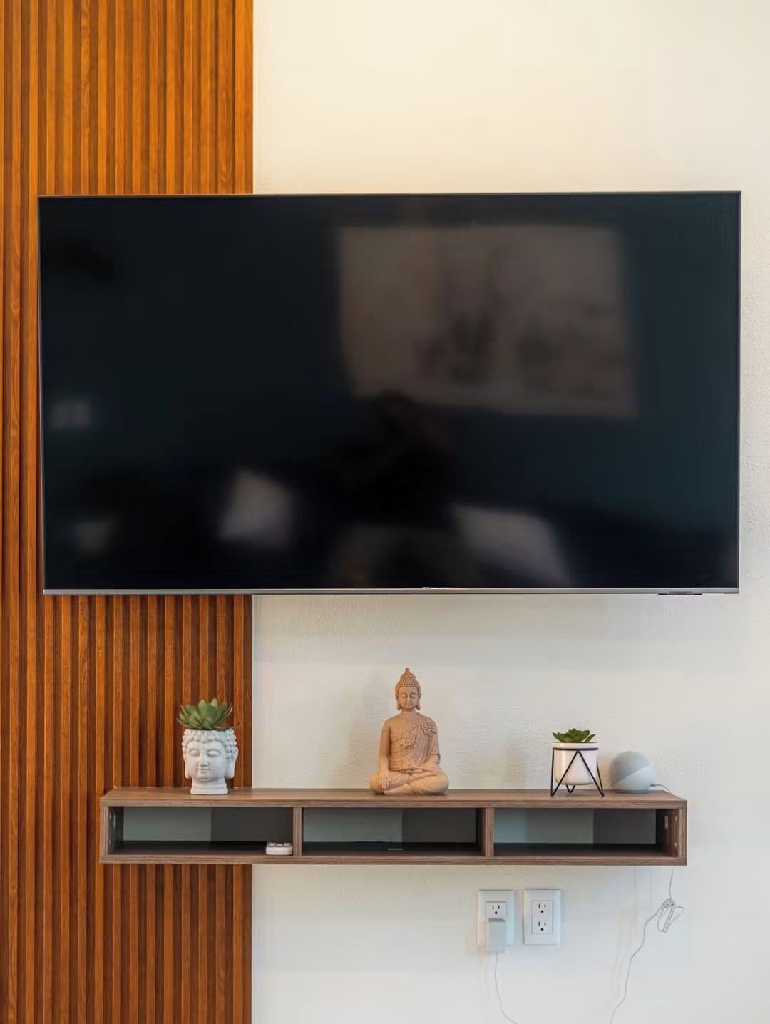 Un téléviseur à écran plat est fixé au mur, lequel est agrémenté d’un panneau en bois. Sous le téléviseur, on trouve une étagère où sont disposés des objets décoratifs, notamment une statue de Bouddha et de petites plantes en pot.
