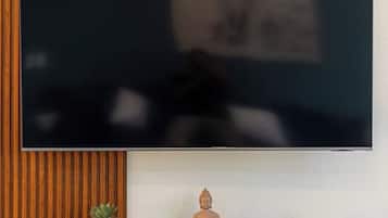 Un téléviseur à écran plat est fixé au mur, lequel est agrémenté d’un panneau en bois. Sous le téléviseur, on trouve une étagère où sont disposés des objets décoratifs, notamment une statue de Bouddha et de petites plantes en pot.